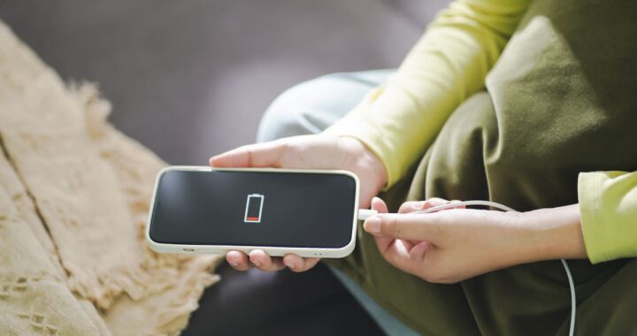 Una mano sostiene un celular con la pantalla de carga encendida, con un cargador y un cable al lado