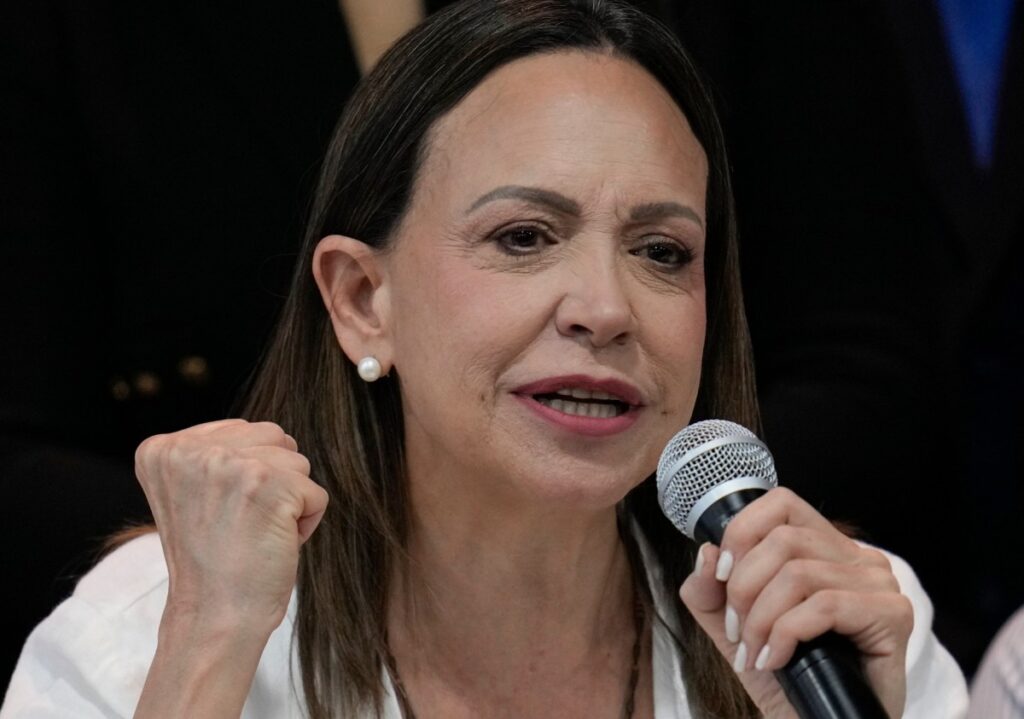María Corina Machado sonriendo mientras habla en un micrófono, con una imagen de Donald Trump detrás.