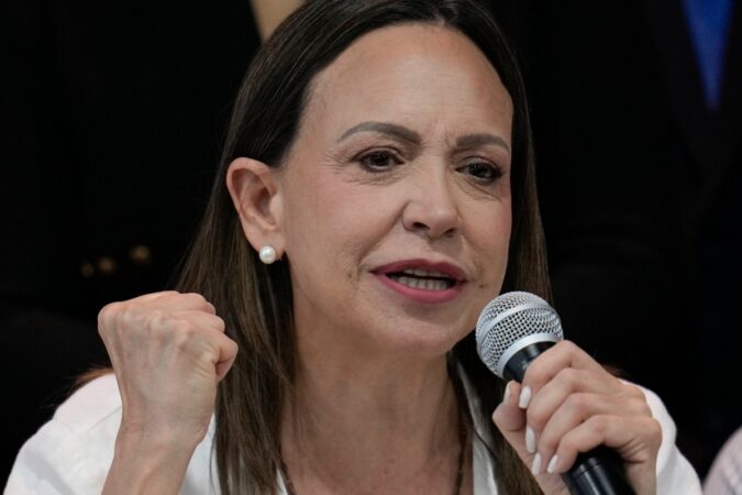 María Corina Machado sonriendo mientras habla en un micrófono, con una imagen de Donald Trump detrás.