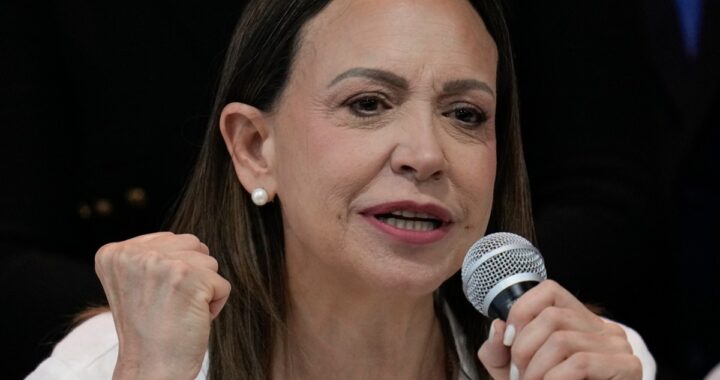 María Corina Machado sonriendo mientras habla en un micrófono, con una imagen de Donald Trump detrás.